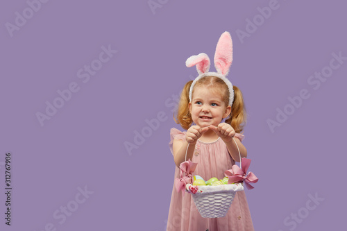 Happy child girl in costume Easter bunny rabbit with ears and a basket with eggs. Little girl on a purple background.