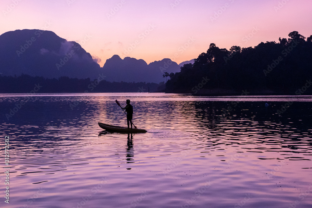Man stand paddling on his kayak at Ratchaprapa dam, or known as Cheow Lan dam, at sunrise Man stand paddling on his kayak at Ratchaprapa dam, or known as Cheow Lan dam, at sunrise