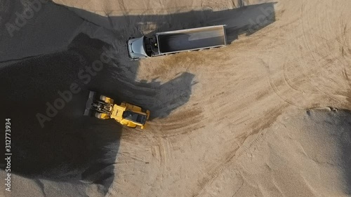 Top aerial view of bulldozer loading stones into empty dump truck in open air quarry.