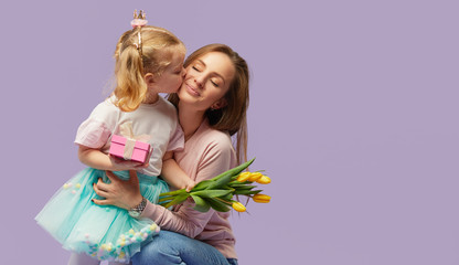 8 march card! Little daughter kisses and hugging her mother with yellow flowers tulip and gift box. Purple studio background. Mothers day concept