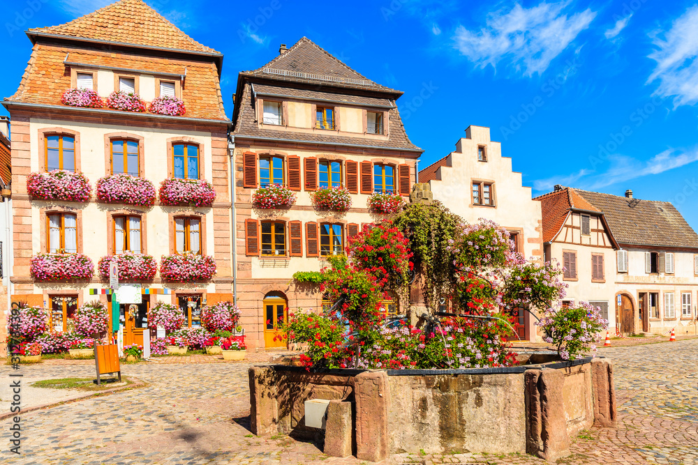 Water fountain and beautiful facades of houses decorated with flowers on square of Bergheim village, Alsace wine route, France