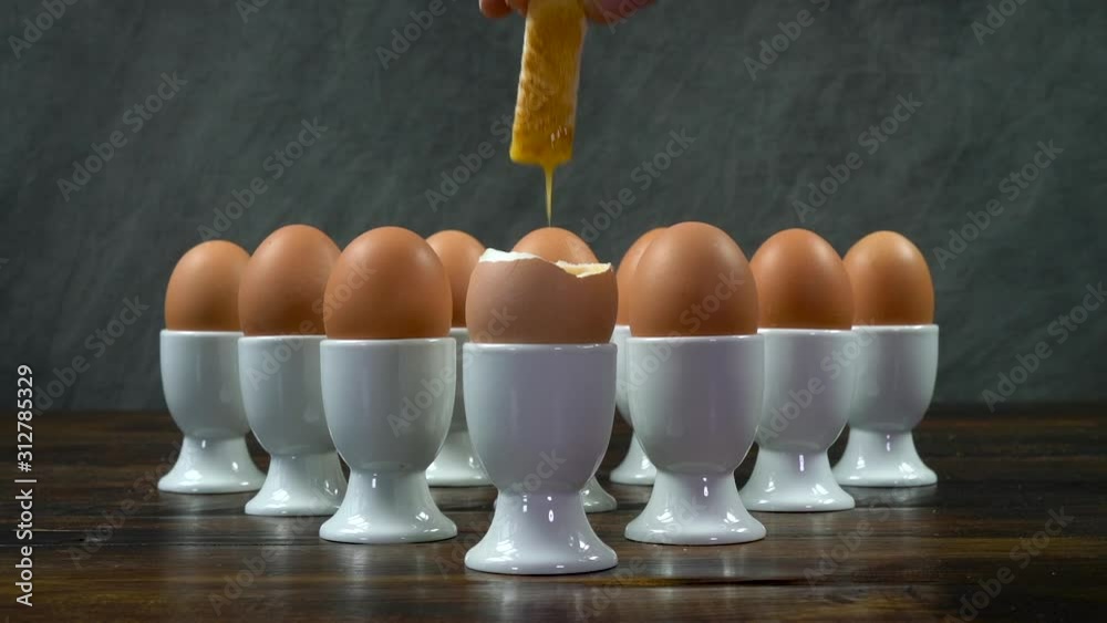 Person dipping a toast soldier into a boiled egg at the front of a group of eggs in white egg cups on a wooden table