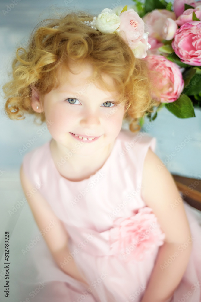 Close up portrait of small 5 years old smiling blonde curly girl in beautiful princess dress with pink flowers on background. Little girl in her birthday waiting for presents. Spring coming. Woman day Close up portrait of small 5 years old smiling blonde curly girl in beautiful princess dress with pink flowers on background. Little girl in her birthday waiting for presents. Spring coming. Woman day