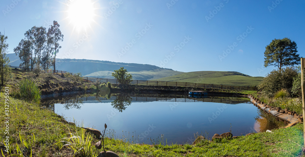 Fototapeta premium Smooth and Calm Dam With The Bright Sun, Blue Sky and Reflective Water, Panorama With A Floating Pontoon in Underberg South Africa