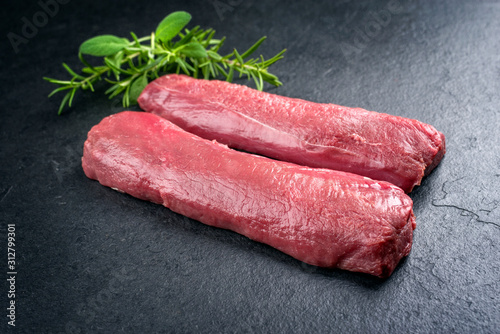 Raw dry aged venison tenderloin fillet steak natural with herbs offered as closeup on a modern design board