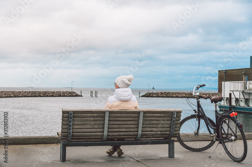 A young Caucasian woman sits with her back on a wooden bench overlooking the Baltic Sea on the seafront in Copenhagen Denmark in winter in cloudy weather. Girl walking gonoskoy bike parked nearby
