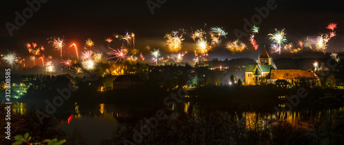 New Year fireworks above the old town of Ratzeburg at night with illuminated cathedral and reflections in the lake, joy for people but panic and fear for animals, panorama view, copy space