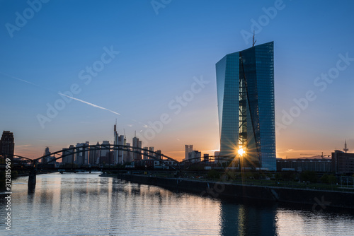 Frankfurt am Main, Germany -May 13th 2019. .European Central Bank (ECB) Tower  and Skyline with a orange-blue Sunsetreflected on the water of the Main River. The sun shines through the ECB towers photo