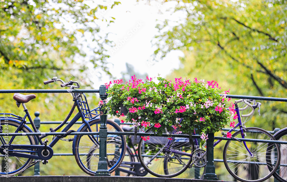 Bikes lined up along a bridge over a canal in Amsterdam, with a large planter of pink flowers. 