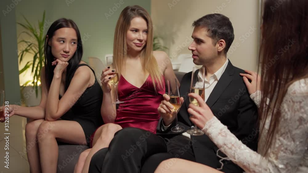 Handsome confident young man talking and flirting with three beautiful women sitting on sofa with flutes of wine at house party Handsome confident young man talking and flirting with three beautiful women sitting on sofa with flutes of wine at house party