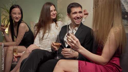 Tracking medium shot of charismatic womanizer man sitting on sofa, talking and flirting with three beautiful young women over sparkling wine at house party photo