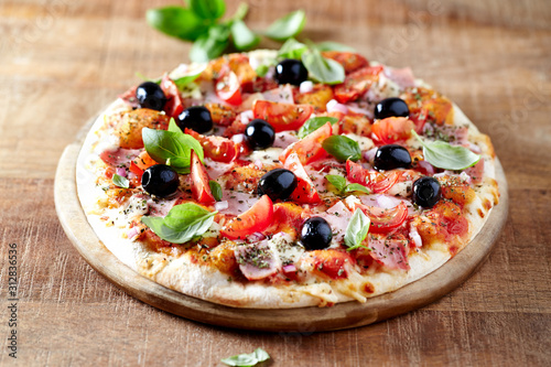 Rustic pizza with ham, cherry tomatoes, black olives and fresh basil on wooden background. 