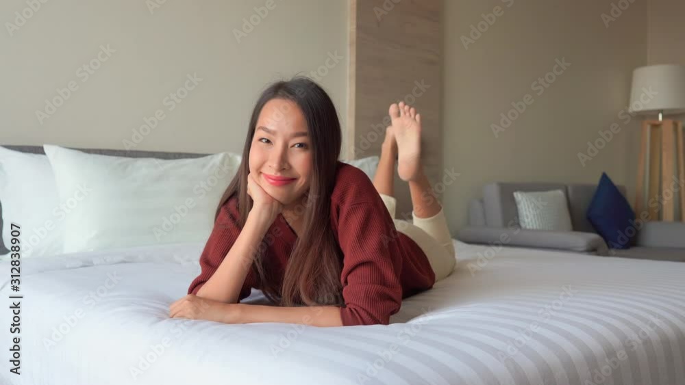 Young adult Asian woman in elegant casual clothing lying on front on freshly made hotel bed rocking with legs. full body shot no shoes. Daylight white beige modern atmosphere and copy space Young adult Asian woman in elegant casual clothing lying on front on freshly made hotel bed rocking with legs. full body shot no shoes. Daylight white beige modern atmosphere and copy space