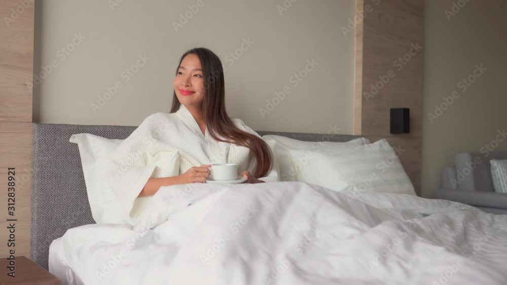 wide shot of young adult Asian woman in white bathrobe in a hotel or hospital rehab double bed holding a cup of tea coffee and looking outside the window smiling. natural daylight, modern interior. wide shot of young adult Asian woman in white bathrobe in a hotel or hospital rehab double bed holding a cup of tea coffee and looking outside the window smiling. natural daylight, modern interior.