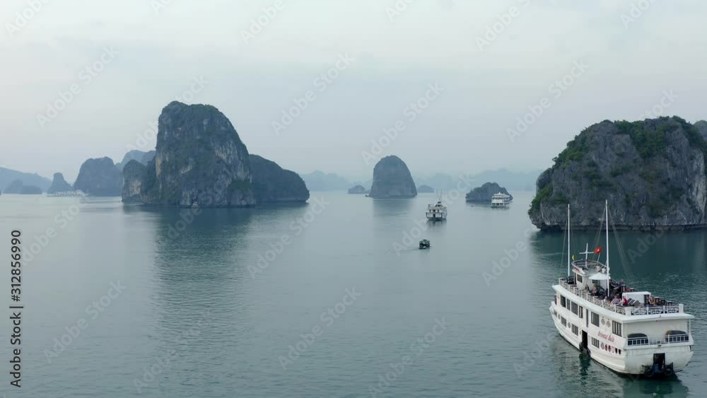 Aerial of cruise ships sitting on the surface of the emerald waters of Ha Long Bay, looking out towards the limestone pillars covered in fog Aerial of cruise ships sitting on the surface of the emerald waters of Ha Long Bay, looking out towards the limestone pillars covered in fog