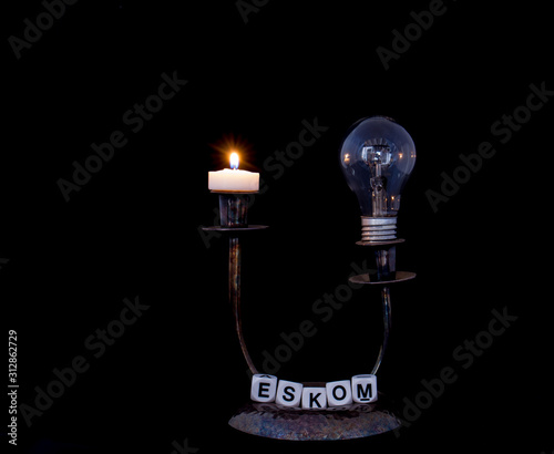 A candle holder with a dead electric bulb and a burning candle with the term Eskom isolated on a black background image with copy space in horizontal format