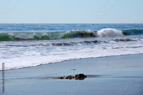 View of the ocean beach in California with surf and sand and a small rock under blue sky on a warm winter day