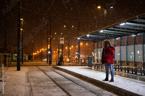 man in red winter coat standing at bus tram stop waiting for public transport