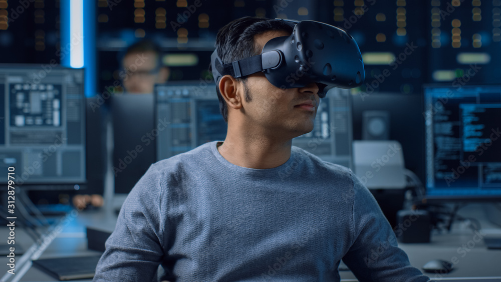 Software Delevoper Wearing Virtual Reality Headset on His Head, Developing and Programming VR Game or Application. In the Background Technology Developing Studio with Computers and Monitors Software Delevoper Wearing Virtual Reality Headset on His Head, Developing and Programming VR Game or Application. In the Background Technology Developing Studio with Computers and Monitors