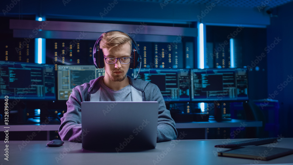 Portrait of Software Developer / Hacker / Gamer Wearing Glasses and Headset Sitting at His Desk and Working / Playing on Laptop. In Background Dark Neon High Tech Environment with Multiple Displays. - obrazy, fototapety, plakaty 