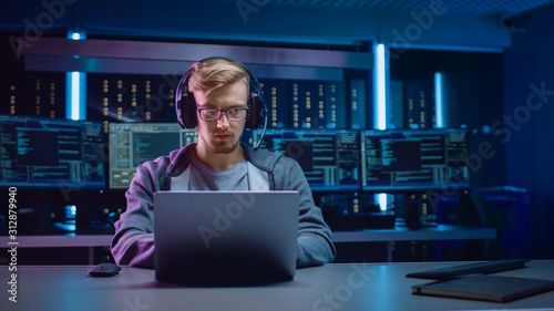 Portrait of Software Developer / Hacker / Gamer Wearing Glasses and Headset Sitting at His Desk and Working / Playing on Laptop. In Background Dark Neon High Tech Environment with Multiple Displays. photo