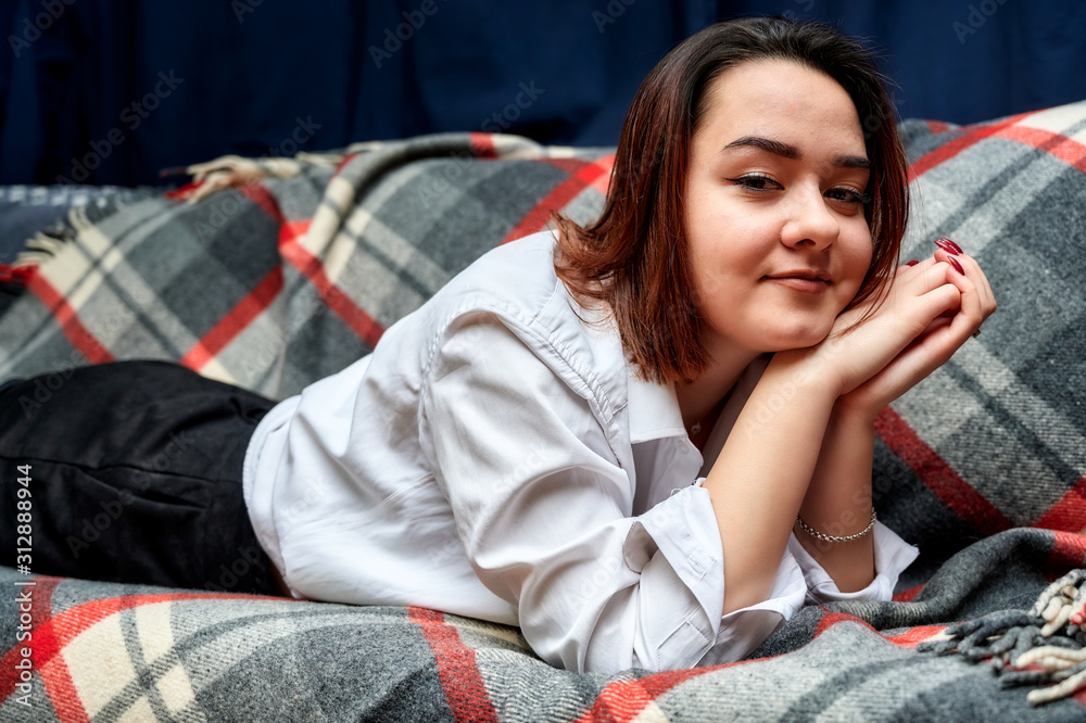 Portrait of a lovely brunette caucasian girl in a white shirt at home on the sofa. Lies and relaxes on a plaid in front of the camera in various poses. Portrait of a lovely brunette caucasian girl in a white shirt at home on the sofa. Lies and relaxes on a plaid in front of the camera in various poses.