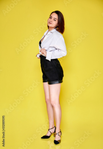 Full-length portrait of a pretty smiling brunette Caucasian girl in a white shirt and black shorts on a yellow background. Standing right in front of the camera in various poses.
