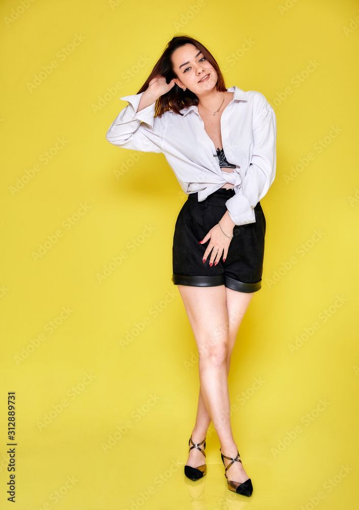Full-length portrait of a pretty smiling brunette Caucasian girl in a white shirt and black shorts on a yellow background. Standing right in front of the camera in various poses. Full-length portrait of a pretty smiling brunette Caucasian girl in a white shirt and black shorts on a yellow background. Standing right in front of the camera in various poses.