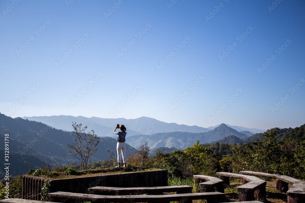 Back view of a woman standing to take pictures of a beautiful mountain view with a smartphone ,Enjoy nature At the viewpoint of Chiang Mai, Thailand
