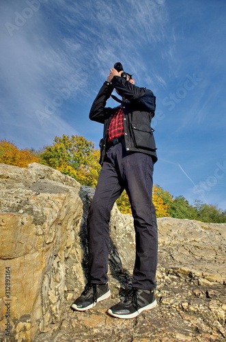 A traveler man standing on the rocks and holding camera over the blue sky background. Vertical frame Side view of a man holding a camera.