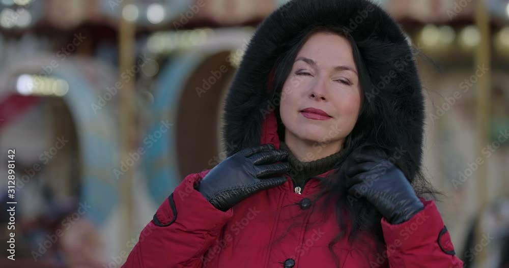 Close-up of middle-aged brunette Caucasian woman in red coat putting on hood. Beautiful positive lady standing on city street and waiting. Leisure, lifestyle, happiness. Cinema 4k ProRes HQ.