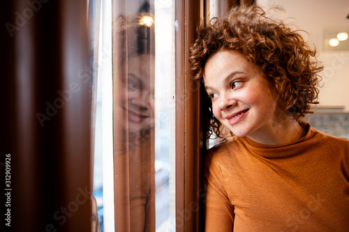 Young woman looking out window, view through glass, close-up. Beautiful woman at the window. Beautiful young brunette woman looking relaxed at home.