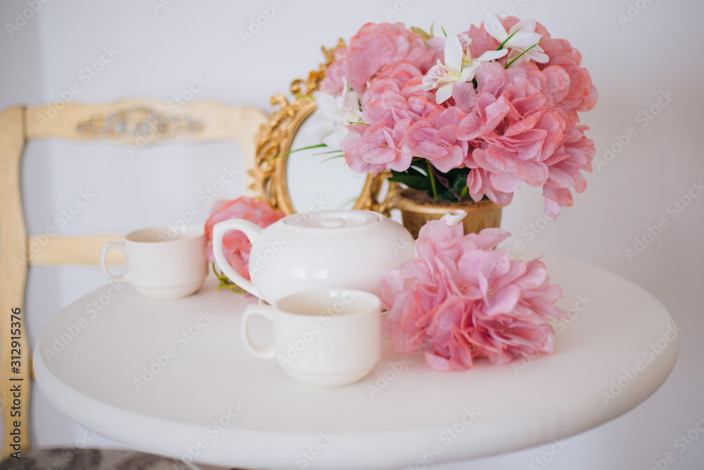 Tea table, mugs and a teapot white teapot with a pink bouquet of flowers in the rococo style. Golden vintage mirror. White pink eltion interior Tea table, mugs and a teapot white teapot with a pink bouquet of flowers in the rococo style. Golden vintage mirror. White pink eltion interior