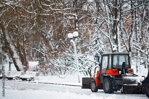 Red small tractor clearing snow in central park, with snow plow and rotating brush. Municipal service removing snow from sidewalk. Road sweeping vehicle with plough brush equipment