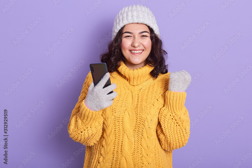 Picture of energetic beautiful female with black curly hair, clenching fist, holding smartphone, smiling sincerely, having pleasant facial expression, being in high spirits. Modern devices concept. Picture of energetic beautiful female with black curly hair, clenching fist, holding smartphone, smiling sincerely, having pleasant facial expression, being in high spirits. Modern devices concept.