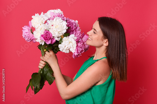 Profile of energeric attractive sweet adorable female looking at bouquet of white and pink flowers with delight, smiling sincerely, standing isolated over red background. Day of lovers concept. photo