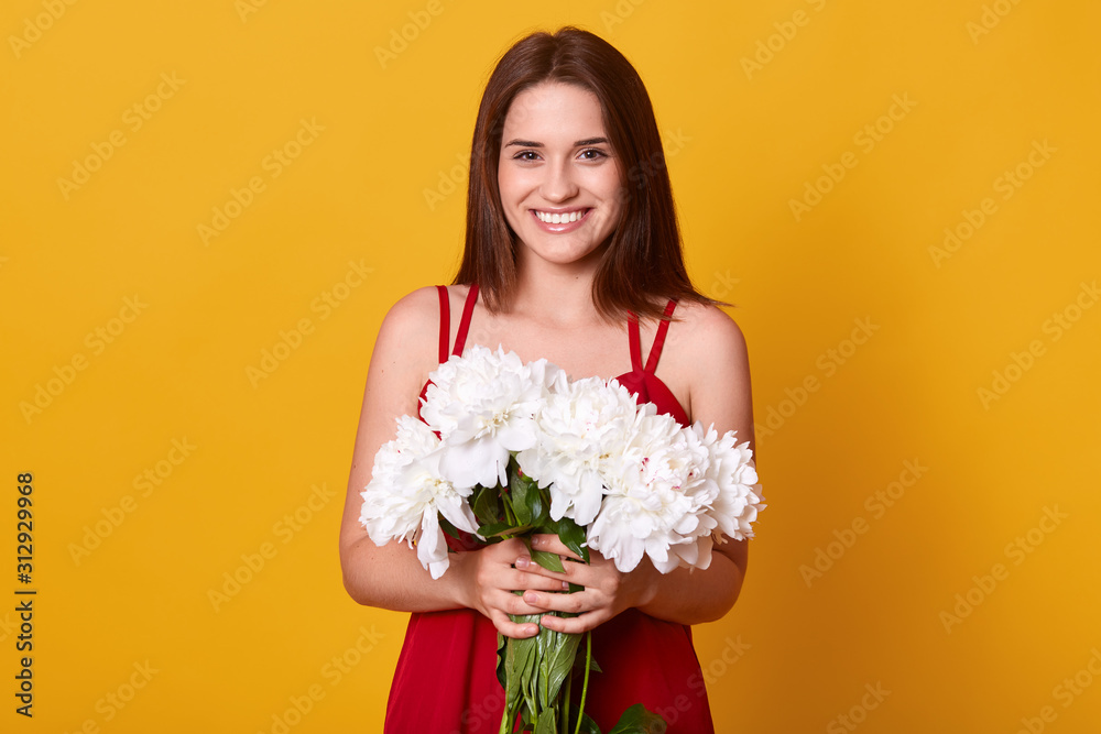 Image of cheerful energetic young lady wearing red dress, having pleasant facial expression, holding white peonies in both hands, smiling sincerely, having black hair. People and emotions concept.