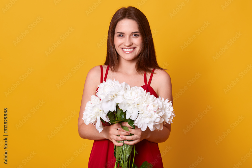 Image of cheerful energetic young lady wearing red dress, having pleasant facial expression, holding white peonies in both hands, smiling sincerely, having black hair. People and emotions concept. Image of cheerful energetic young lady wearing red dress, having pleasant facial expression, holding white peonies in both hands, smiling sincerely, having black hair. People and emotions concept.