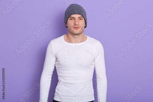 Closeup portrait of handsome magnetic man standing isolated over lilac background in studio, wearing grey hat and white sweatshirt, looking directly at camera, keeping strong fit. Cold times concept. photo