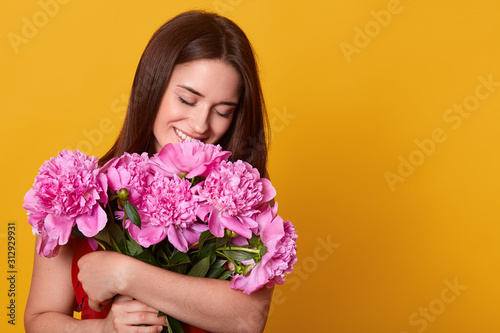 Indoor studio shot of tender charming young female having pleasant facial expression, smelling pink peonies, getting pleasure of presents, being in high spirits. Copyspace for advertisement.