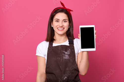 Indoor studio shot of happy attractive energetic female smiling sincerely, holding switched off tablet in one hand, looking directly at camera, having pleasant facial expression. Digital concept.