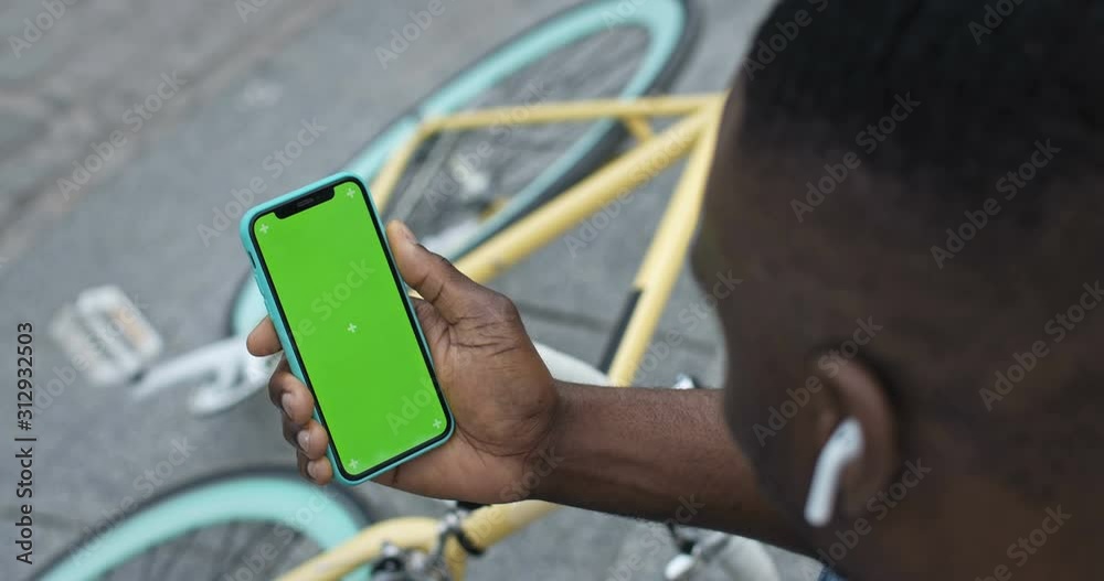 Lviv, Ukraine - August 09, 2019: Close up of African American Man in Earphones Holding his Smartphone Touching Green Screen and Looking to it while Sitting on Steps near Bike at the City Background.