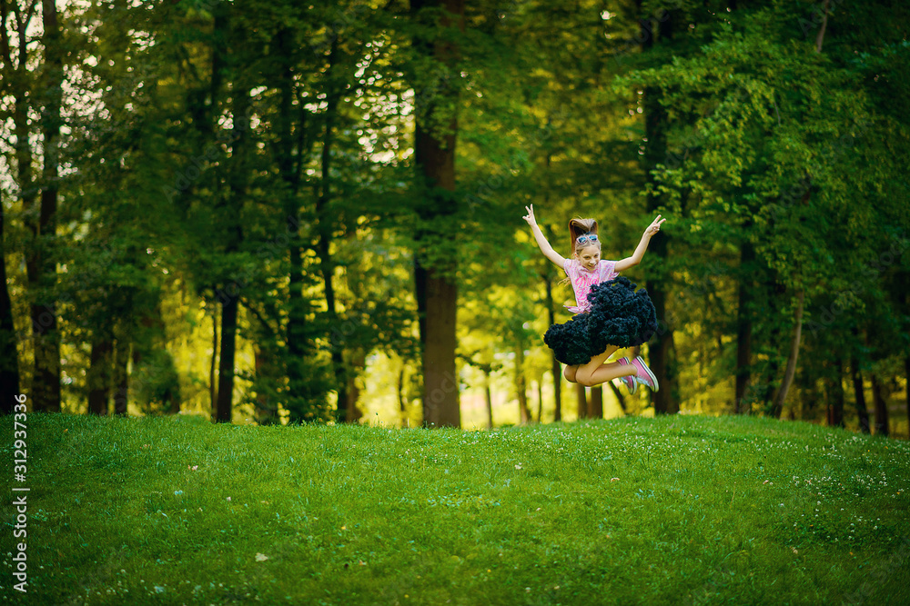 A beautiful girl in a fluffy black skirt tutu joyfully jumps on a green lawn among the trees. The concept of happy childhood.