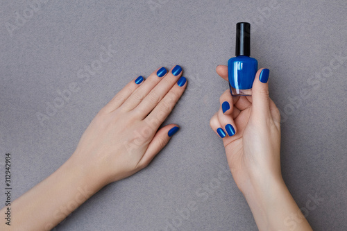 Female hands with manicure in trendy classic blue color holding a bottle of nail polish on the grey background