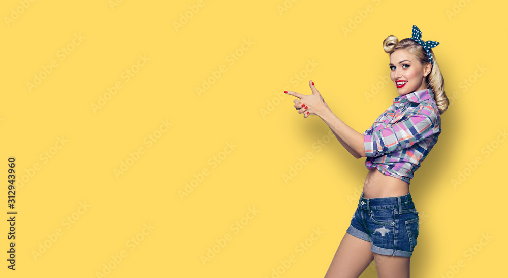 Happy young woman pointing. Excited girl in pin up style, showing something or copy space for advertise slogan or text. Retro fashion and vintage hairdo concept. Yellow color background. Happy young woman pointing. Excited girl in pin up style, showing something or copy space for advertise slogan or text. Retro fashion and vintage hairdo concept. Yellow color background.