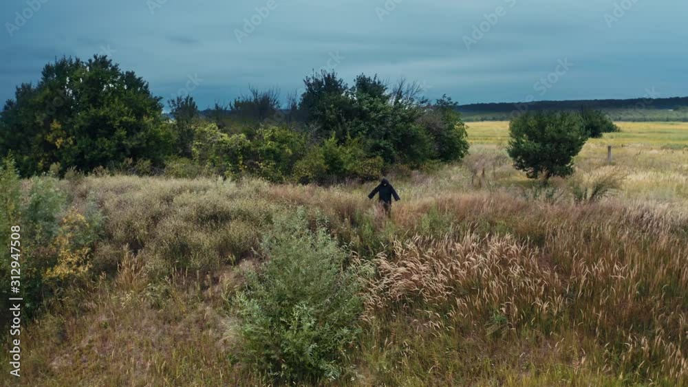 Devil in black costume in nature. Field in summer and dark figure in black robe with outstretched arms standing. Horrorful ghost outdoors. Devil in black costume in nature. Field in summer and dark figure in black robe with outstretched arms standing. Horrorful ghost outdoors.