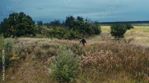 Devil in black costume in nature. Field in summer and dark figure in black robe with outstretched arms standing. Horrorful ghost outdoors. photo
