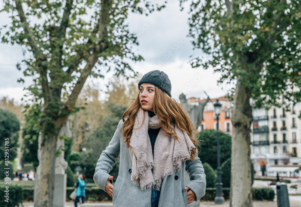Fototapeta premium Young blonde European woman, carries her hands in the pockets of her coat, while walking through a park on a cold day