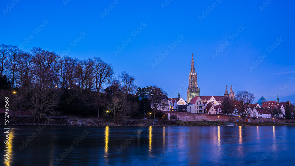 Germany, Ulm city skyline, cityscape and minster cathedral behind water of danube river in beautiful blue hour atmosphere by night after sunset