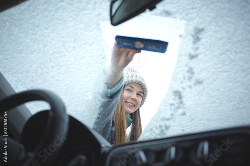 Smiling girl cleaning frozen windshield of a car from snow.